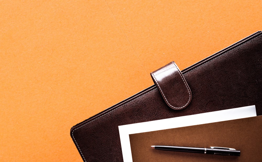 A brown leather case, a notepad and a black pen against an orange background 
