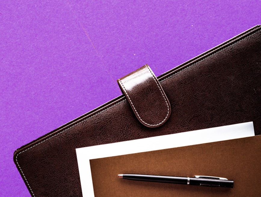 A brown leather case, a notepad and a black pen against a purple background