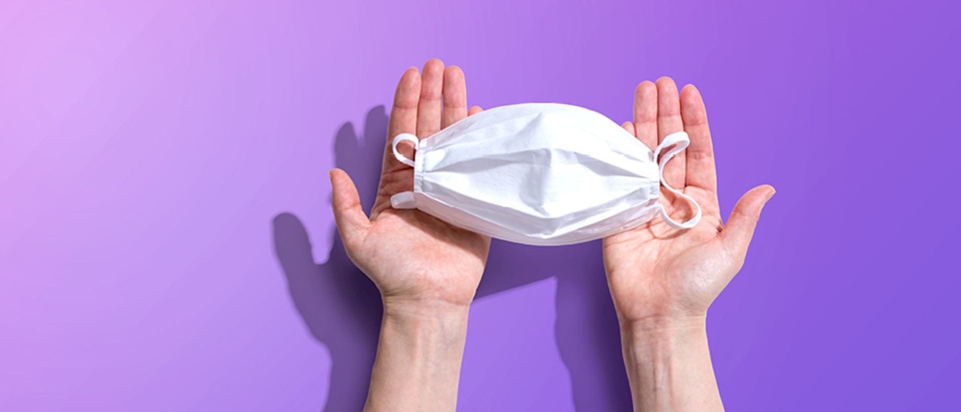 Image of a white surgical mask in a pair of hands on a purple background