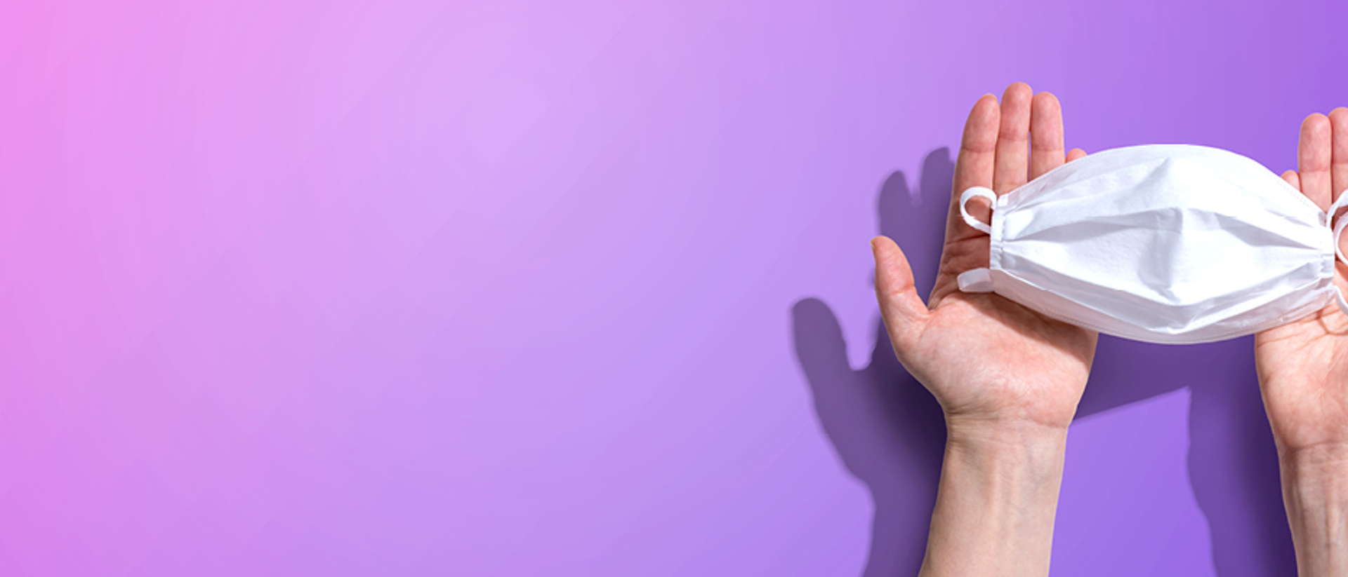 Image of a white surgical mask in a pair of hands on a purple background