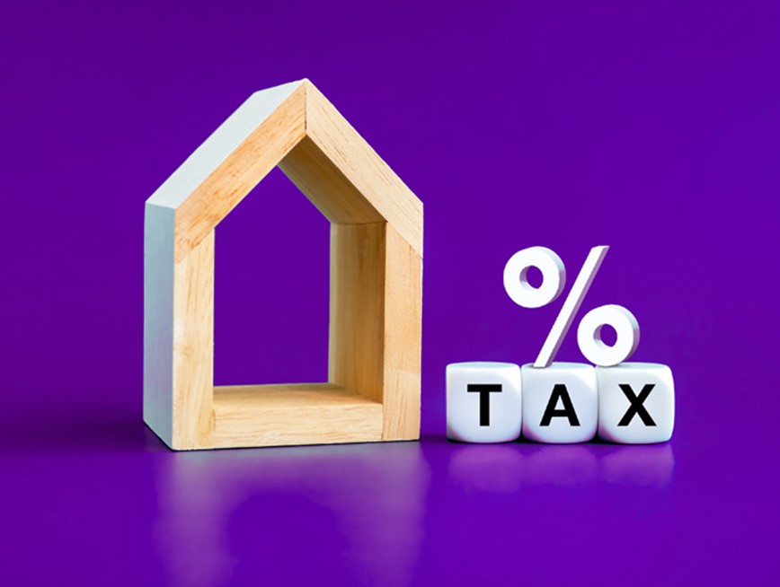 A wooden block shaped as a house, with a percentage symbol on top of dice spelling out TAX, all against a purple background.