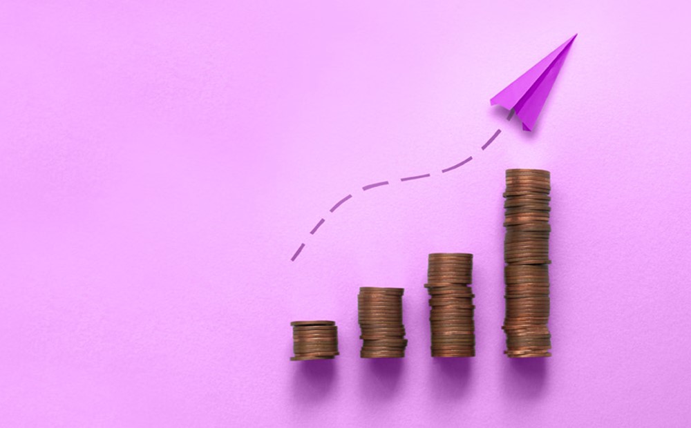 Image of a purple paper airplane flying over stacks of coins against a purple background
