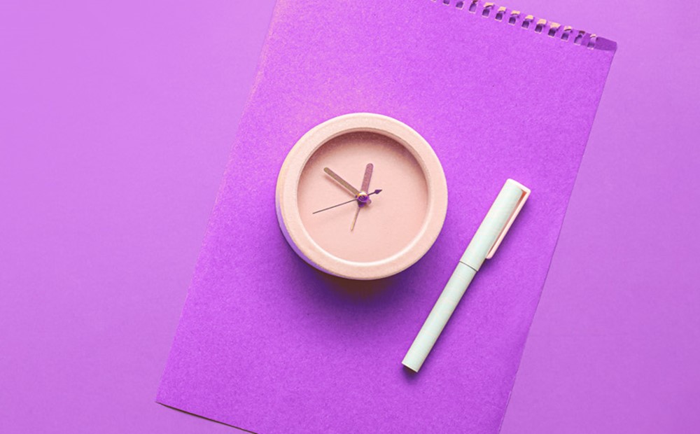 A peach coloured clock and a white pen on top of a purple folder against a purple background