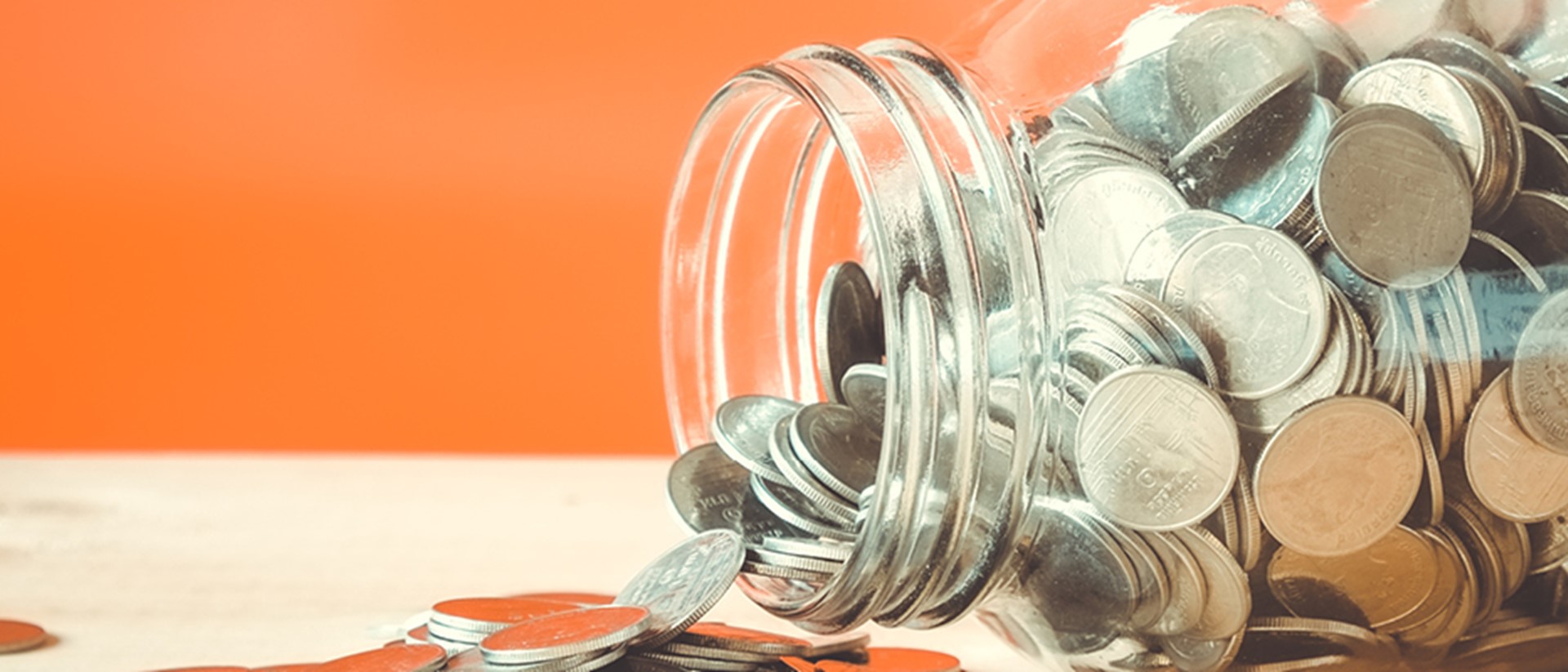Image of coins flowing our of a glass jar on it's side on an orange background