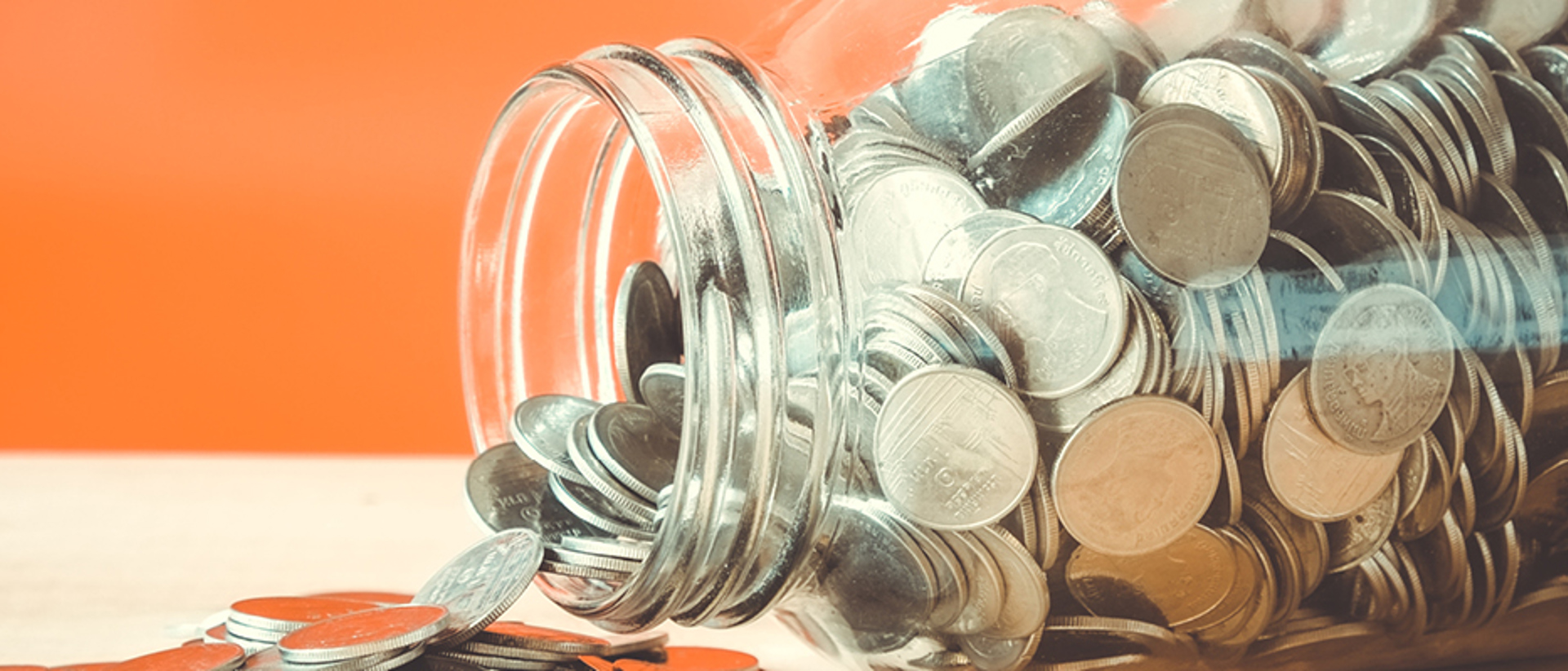 Image of coins flowing our of a glass jar on it's side on an orange background